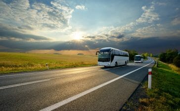 Two white buses traveling on the asphalt road in rural landscape at sunset with dramatic clouds